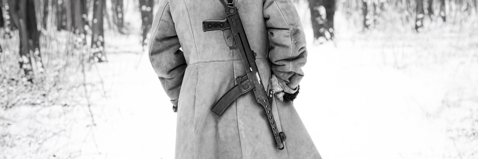 A soldier seen from behind, greatcoat and slung rifle, walking into a snowy forest